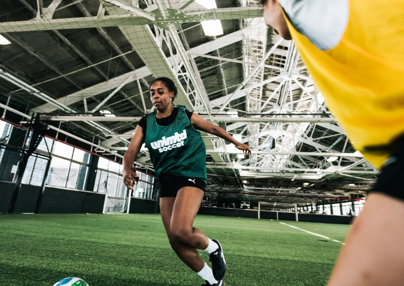 Joueuse de soccer en maillot vert dribblant un ballon coloré sur un terrain de gazon intérieur avec une autre joueuse en