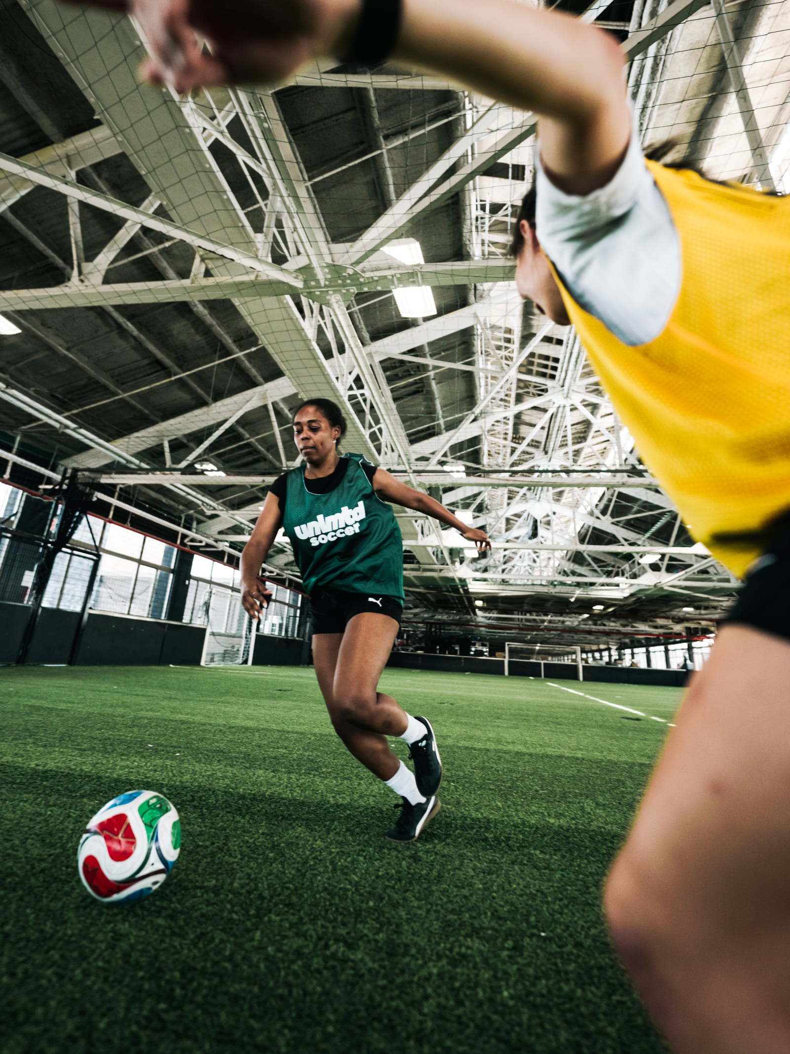 Female soccer player in green jersey dribbling a colorful ball on an indoor turf field with another player in yellow nearby