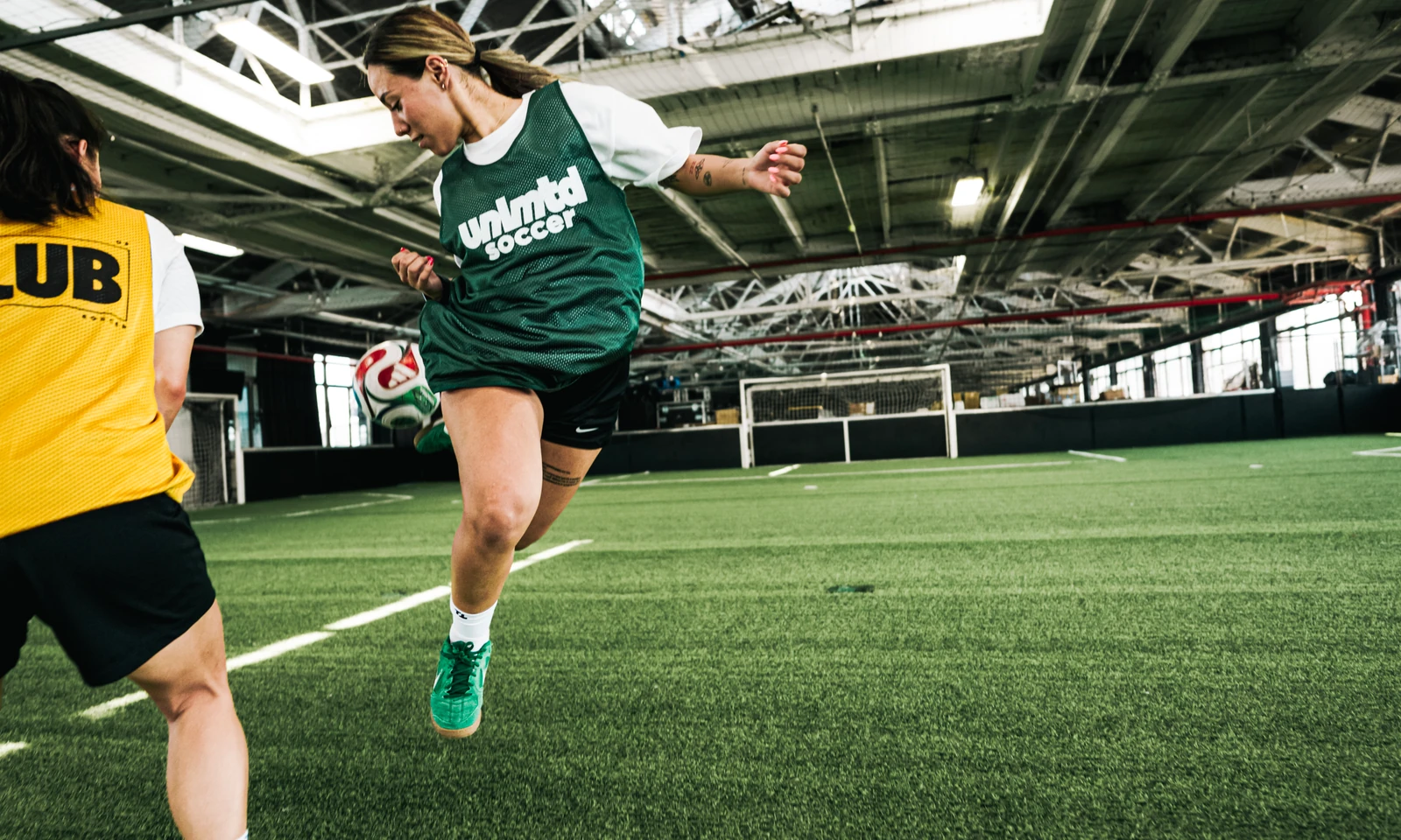 Female soccer player in green jersey kicking ball during indoor training session with teammate in yellow jersey