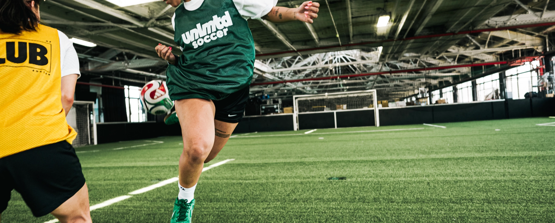 Female soccer player in green jersey kicking ball during indoor training session with teammate in yellow jersey
