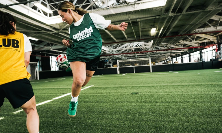Female soccer player in green jersey kicking ball during indoor training session with teammate in yellow jersey