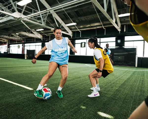 Deux joueuses de soccer en maillots bleu clair et jaune s'entraînent sur un terrain de turf intérieur avec un ballon de