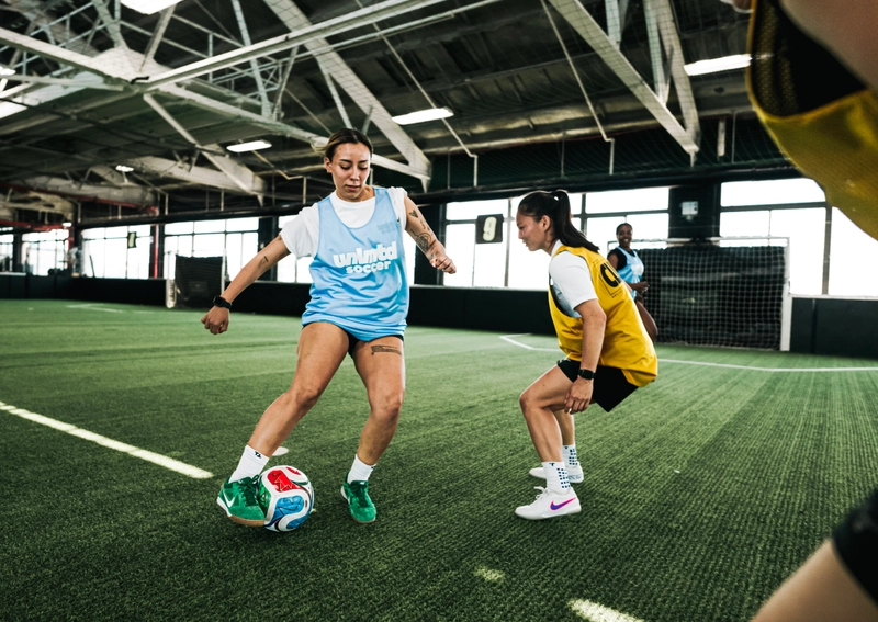 Deux joueuses de soccer en maillots bleu clair et jaune s'entraînent sur un terrain de turf intérieur avec un ballon de