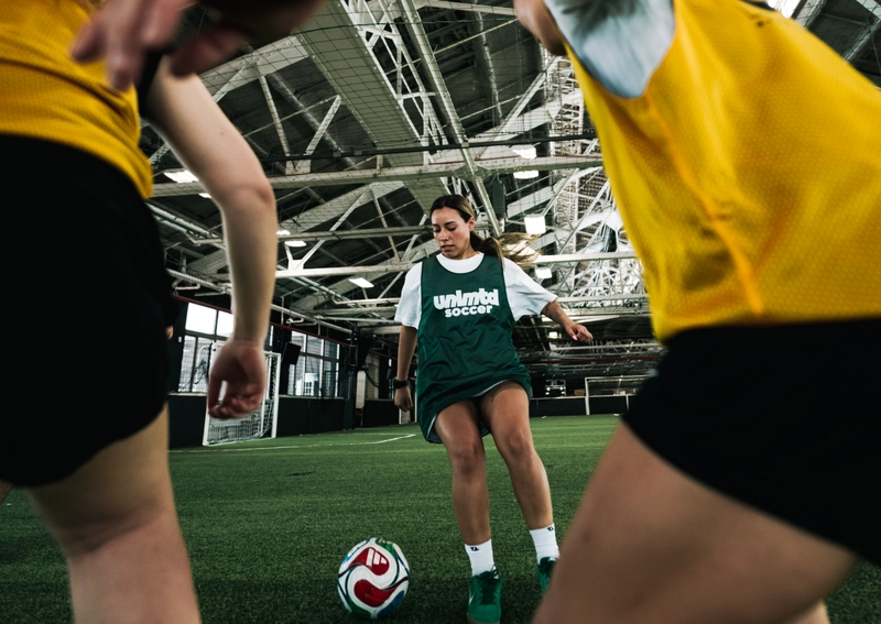 Des joueuses de soccer en gilets jaunes et un maillot vert avec la marque unlimited soccer s'entraînent en intérieur sur une