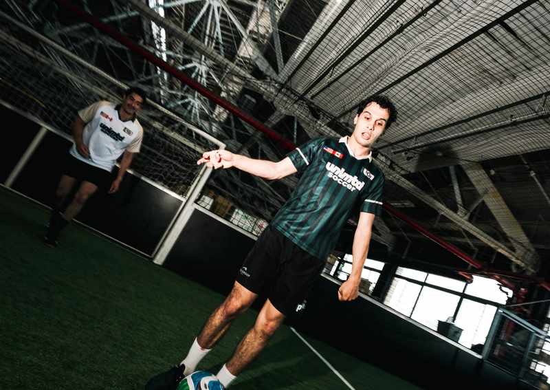 Un joueur de soccer en maillot vert équilibre un ballon coloré sur un terrain de gazon intérieur, avec un autre joueur en