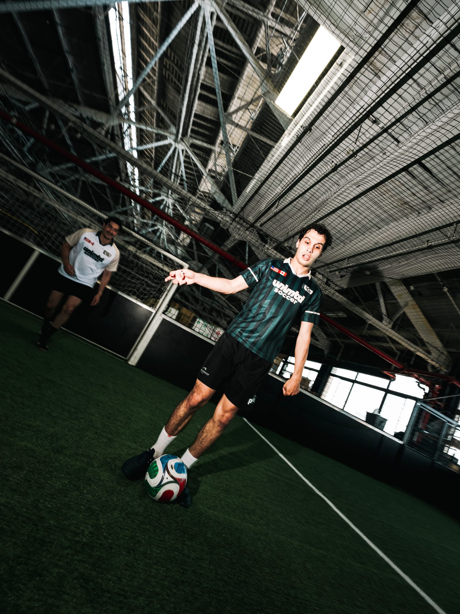 A soccer player in a green jersey balances a colorful ball on an indoor turf field, with another player in the background