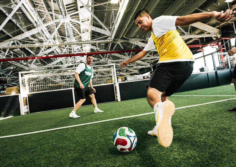 Un joueur en gilet jaune frappe un ballon de soccer sur un terrain de gazon artificiel intérieur, avec un autre joueur en