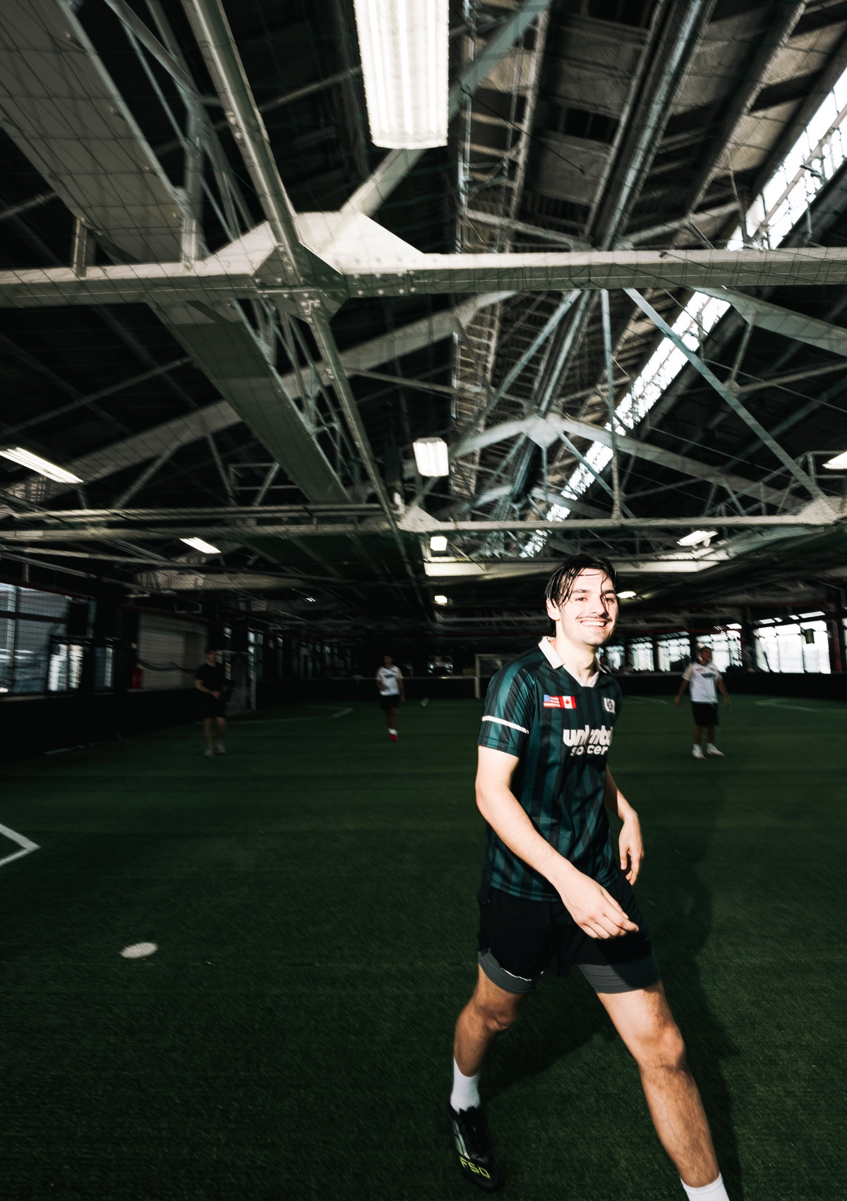 Un joueur souriant en maillot rayé vert et noir marche sur un terrain de soccer intérieur avec gazon artificiel et poutres