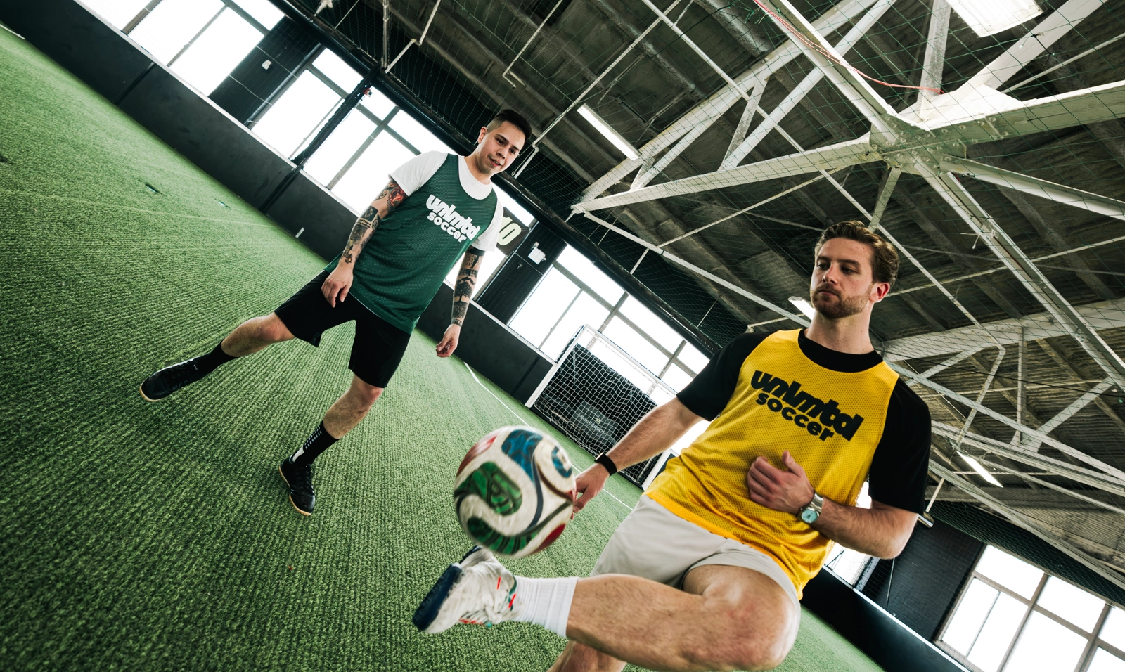 Deux hommes en maillots Unlmtl Soccer s'entraînent sur un terrain de gazon artificiel intérieur avec des poutres de plafond