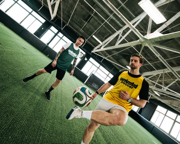 Deux hommes en maillots Unlmtl Soccer s'entraînent sur un terrain de gazon artificiel intérieur avec des poutres de plafond