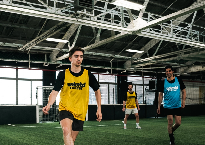Trois joueurs en maillots jaunes et bleus unlmtd soccer s'entraînent sur un terrain de turf intérieur avec des poutres de
