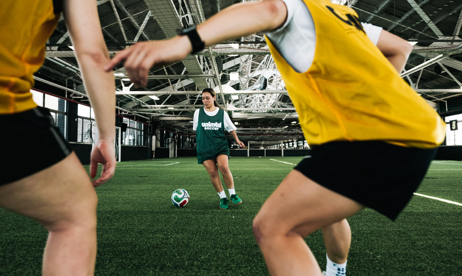Jeunes joueurs de soccer en gilets jaunes et maillot vert s'entraînent sur un terrain de gazon artificiel en intérieur avec