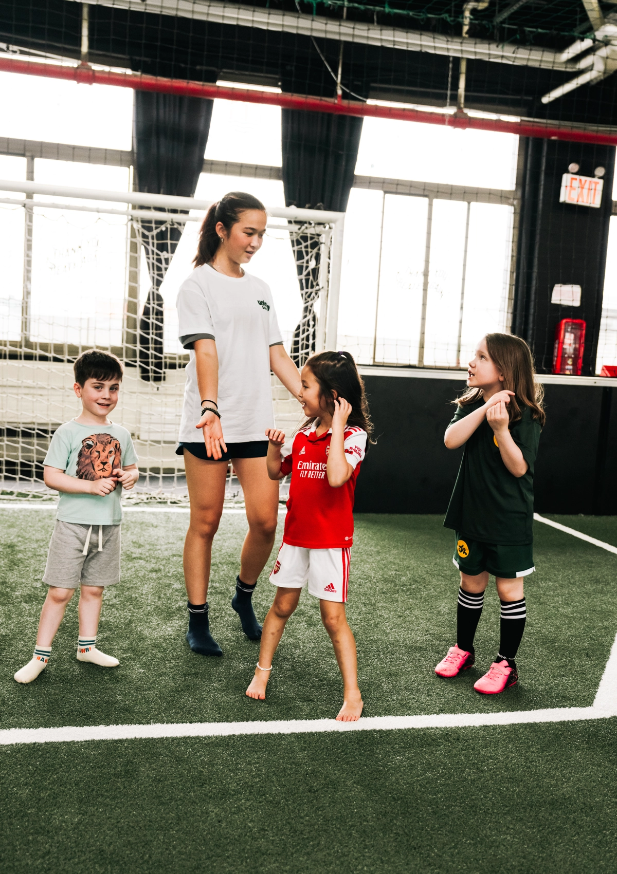 A coach interacts with three children on an indoor soccer field, with a goal net and large windows in the background.