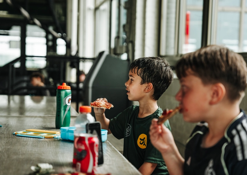 Two young boys in soccer jerseys enjoy pizza at a modern indoor cafe table with drinks and snacks, likely during a break at