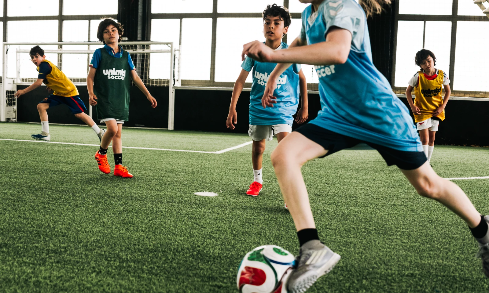 Young players in blue and yellow jerseys actively engaged in an indoor soccer training session on artificial turf, with one