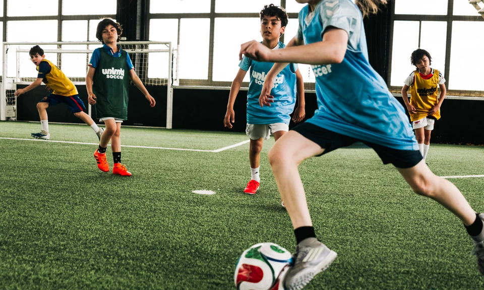 Young players in blue and yellow jerseys actively engaged in an indoor soccer training session on artificial turf, with one