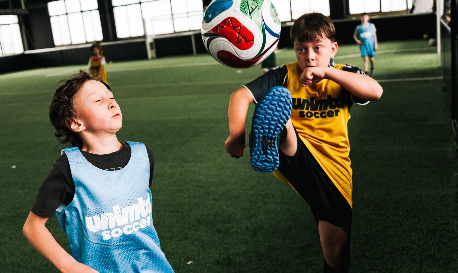 Deux enfants en maillots bleu et jaune jouent avec un ballon de soccer sur une pelouse artificielle en intérieur dans un