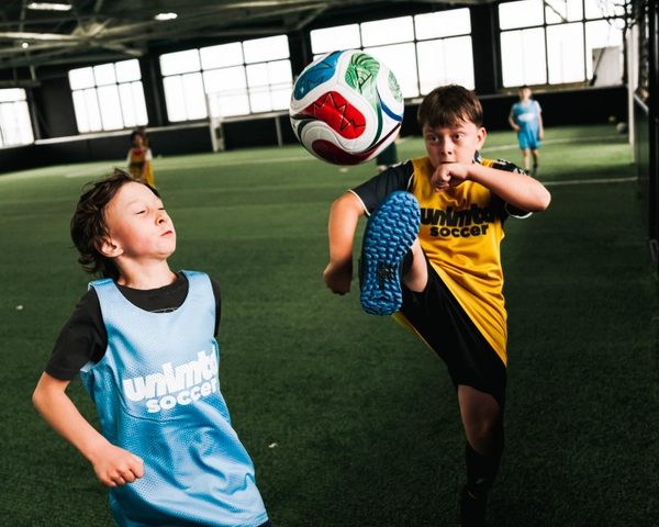 Deux enfants en maillots bleu et jaune jouent avec un ballon de soccer sur une pelouse artificielle en intérieur dans un