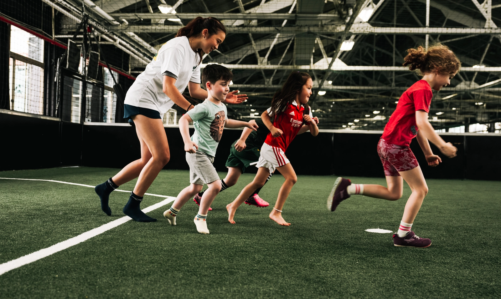 Children and a coach sprinting during a soccer training session on an indoor artificial turf field with exposed ceiling