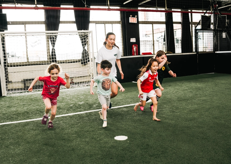 Children running during a soccer training session on an indoor field with a coach and goal net in the background