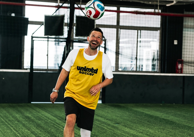 Un homme en gilet jaune unlimited soccer jongle un ballon de soccer coloré sur une pelouse artificielle intérieure avec du