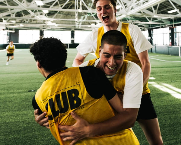 Trois joueurs de soccer en maillots jaunes célébrant un but sur un terrain de gazon intérieur avec un haut plafond et des