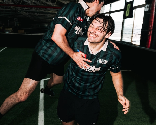 Deux joueurs de soccer en maillots verts à carreaux célébrant sur un terrain de gazon intérieur, l'un sautant sur le dos de