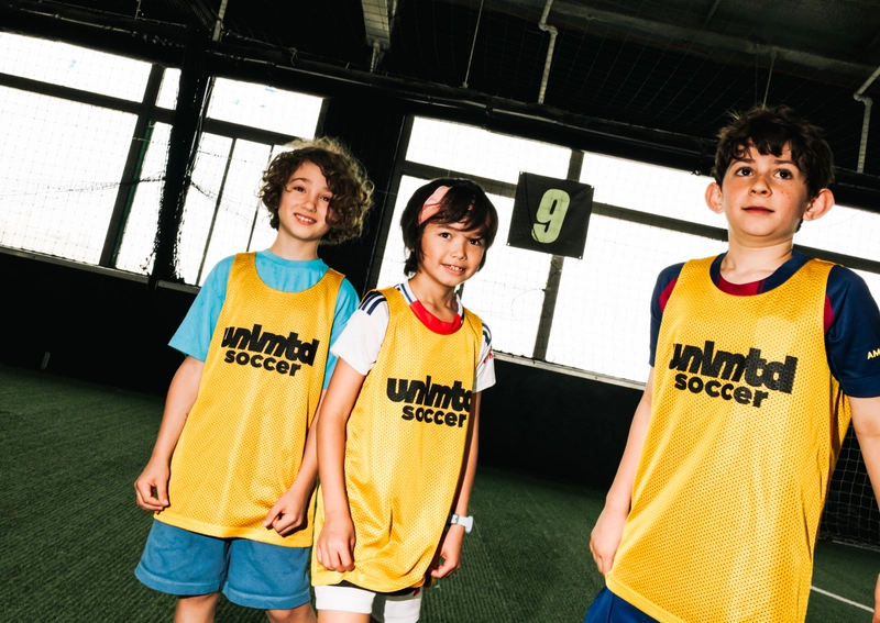 Trois jeunes joueurs de soccer en gilets jaunes unlmtd soccer debout sur un terrain intérieur avec des ballons de soccer