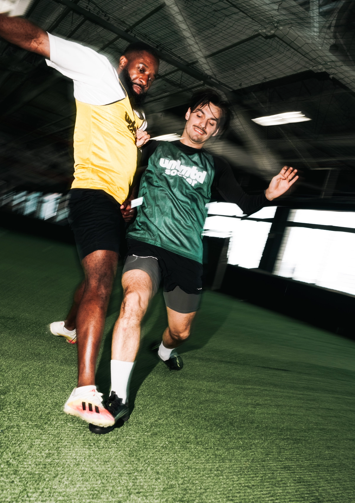 Two soccer players in action on an indoor field, one in a yellow jersey and the other in a green "unlmtdsoccer" jersey