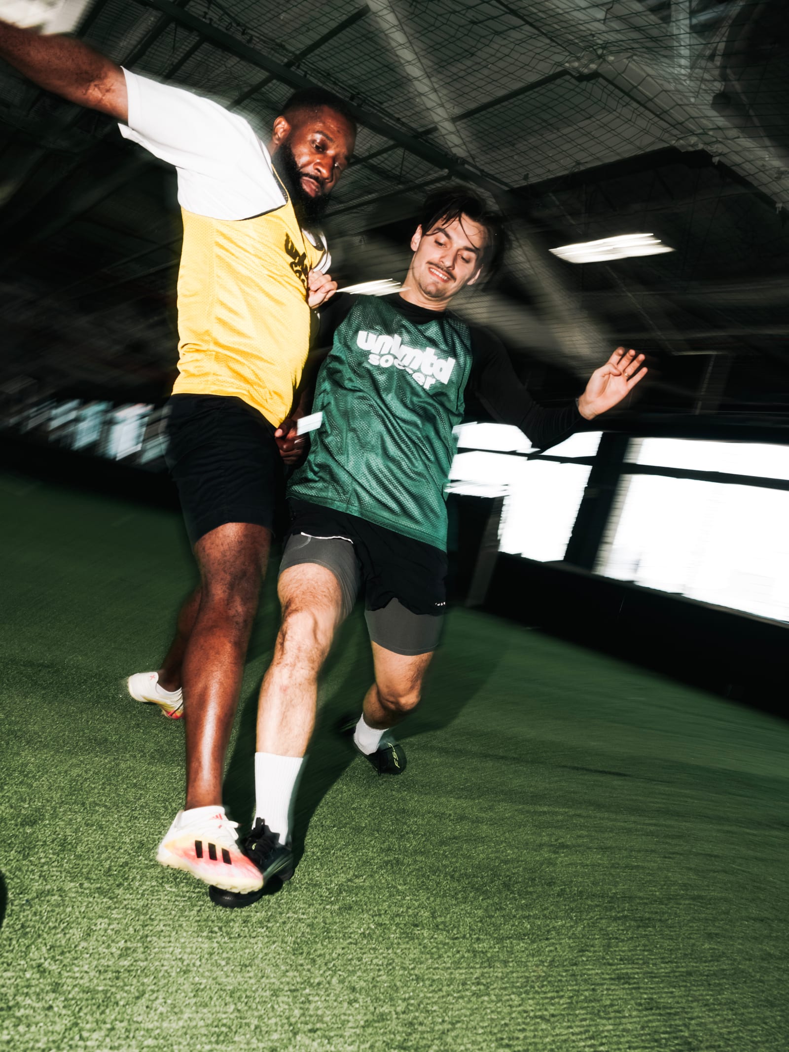 Deux joueurs de soccer en action sur un terrain intérieur, l'un en maillot jaune et l'autre en maillot vert "unlmtdsoccer"