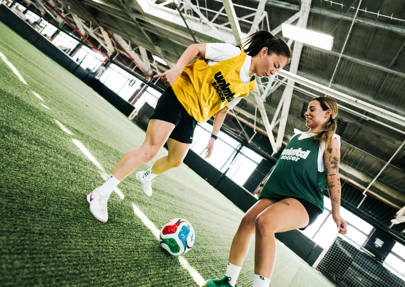 Two women in sports jerseys play soccer on an indoor turf field with a colorful ball, surrounded by industrial-style