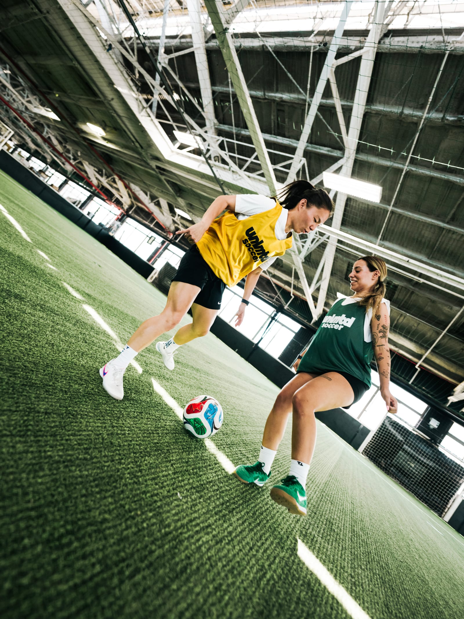 Deux femmes en maillots de sport jouent au football sur une pelouse artificielle intérieure avec un ballon coloré, entourées