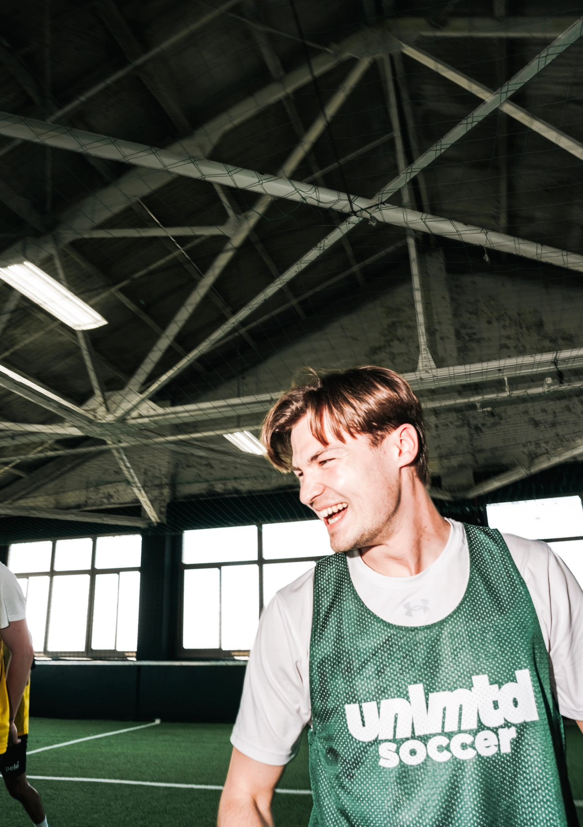 Un joueur de soccer souriant, vêtu d'un maillot vert "unlmtd soccer", se trouve sur un terrain de gazon artificiel intérieur