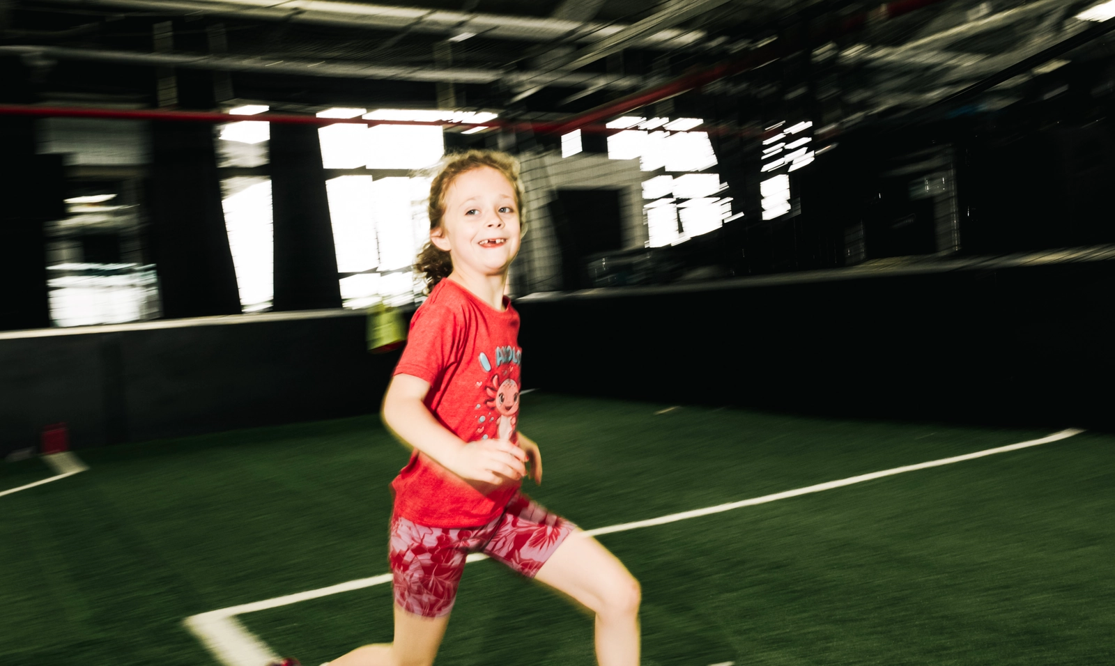 Une jeune fille en T-shirt rouge et short à motif court avec énergie sur un terrain de soccer intérieur avec gazon