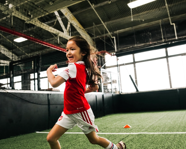 Une jeune fille en maillot de football rouge et blanc court avec énergie sur une pelouse artificielle intérieure, avec un