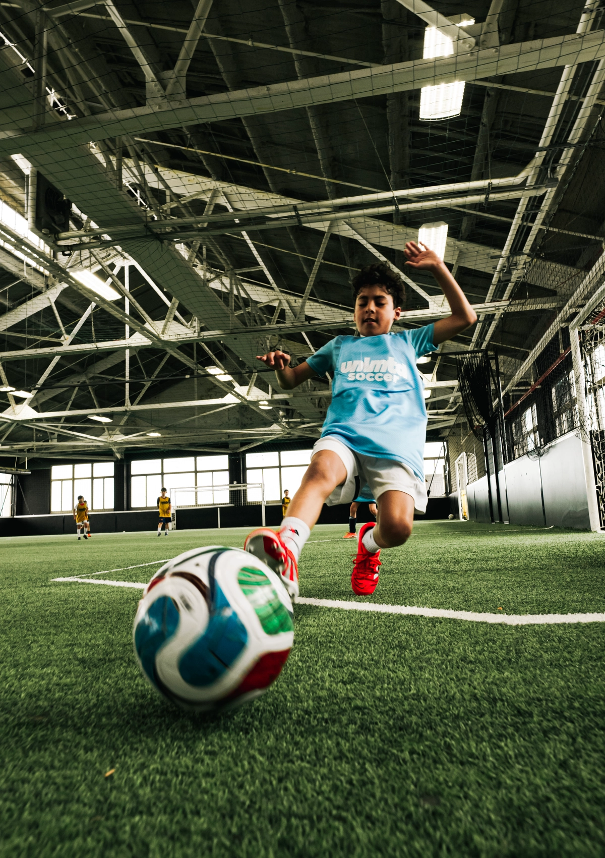 Un jeune joueur en maillot bleu clair et chaussures rouges frappe un ballon de soccer coloré sur un terrain de gazon