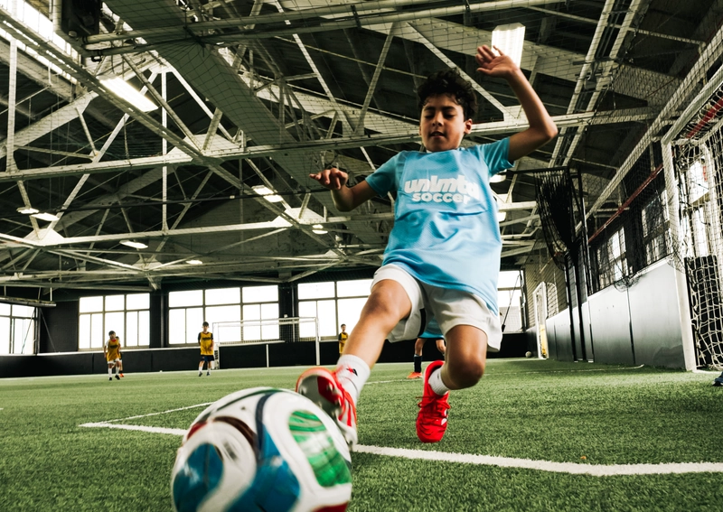 A young player in a light blue shirt and red shoes kicks a colorful soccer ball on an indoor turf field with a goal and