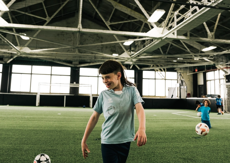 Un jeune joueur de soccer sourit tandis qu'il marche sur un terrain de gazon artificiel intérieur avec des ballons de soccer