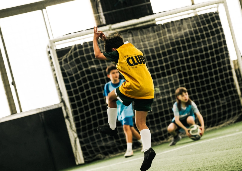 Un jeune joueur de soccer en maillot jaune saute avec les bras levés près d'un filet de but, tandis que des coéquipiers en