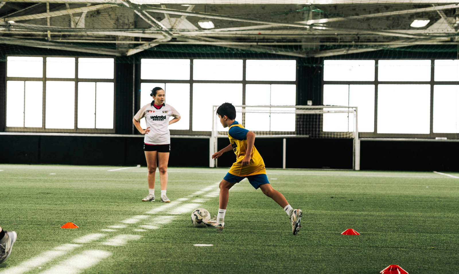 A young soccer player in a yellow jersey dribbles a ball around cones during a training session on an indoor turf field