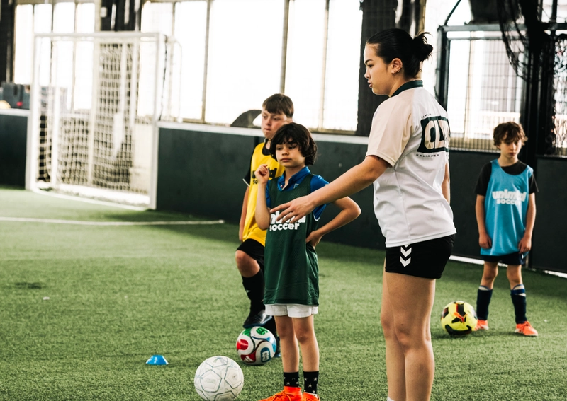 A youth soccer coach instructing young players during a training session on an indoor turf field with soccer balls and cones